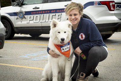 Detective Tara Hurless kneels beside Kirby, a fluffy white dog, in front of a University Police vehicle. The dog is wearing a bandana around his neck that says "Hello my name is Kirby."