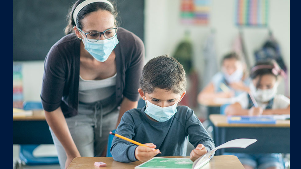 student teacher watches a young boy doing schoolwork as both wear masks