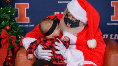 person presenting as Santa Claus holds a baby on his/her/their lap in front of a Fighting Illini backdrop