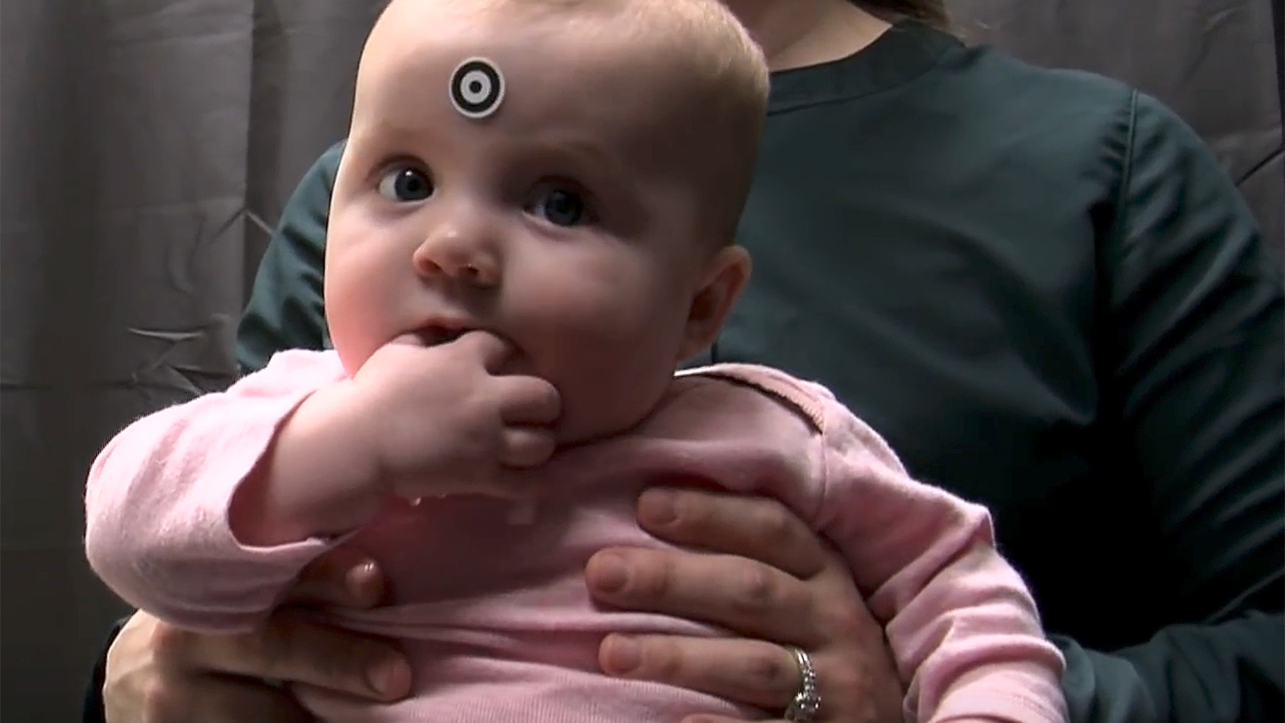 Photo of an infant seated on her mother’s lap. The infant has a sticker on her forehead that allows the eye-tracking instrument to orient to her eyes.  Photo by Steven Drake