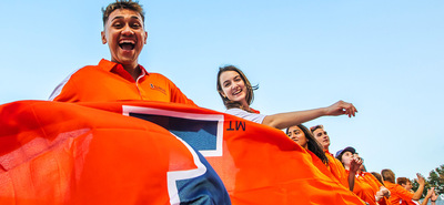 grinning male student dressed in orange waiving a big orange Illini flag in a line of other super happy students similarly attired in Illini gear all set against a clear blue sky.
