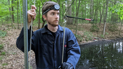 The author helps his colleagues install mist nets around a pond. ALT TEXT: Photo of the author standing near a vertical pole used to secure the nets. He grasps a cord used to tighten the nets in place.  Photo by Elizabeth Beilke