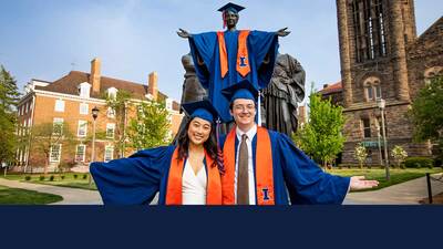 two students in caps and gowns pose in front of the Alma Mater statue, which is also wearing cap and gown