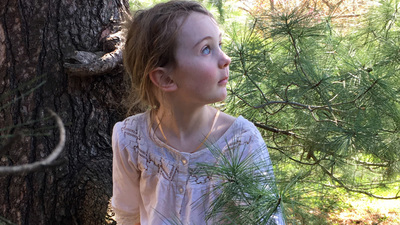 young girl looks around from her vantage point at the base of a pine tree