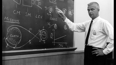 John Houbolt stands in front of chalk board explanation of Lunar-Orbit Rendezvous