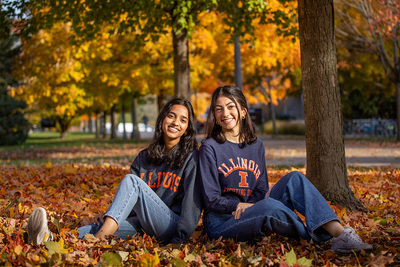 Two smiling students sitting under a tree on the U. of I. quad in the autumn surrounded by fallen leaves