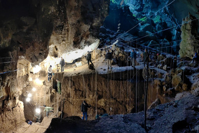 Image of the interior of the cave and the massive trench with people standing at different levels and looking into the trench. The cave is dark and you can see the grid of guidelines used to plot the location of items found in the dig. There are bright worklights overhead.