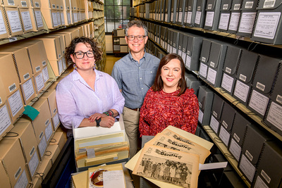Photo of Jenny Davis, Christopher Prom and Bethany Anderson standing between library stacks filled with boxes, with materials including newspapers spread in front of them.