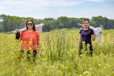 Holly Tuten and graduate student Erica Hernandez stand in a prairie with drag cloths attached to poles over their shoulders. They are smiling and looking at the camera.