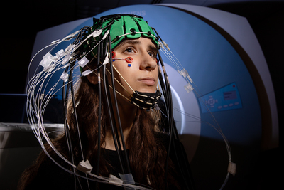 Photo of a young woman inside an MRI suite wearing an imaging cap with many sensors attached.