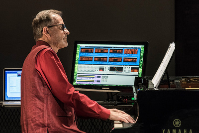 Photo of David Rosenboom at a piano keyboard with a computer screen next to him.