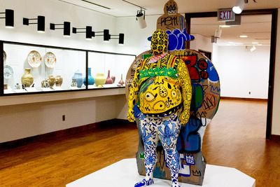 Photo of a life-sized cast of a man's body, standing against the sheared-off side of a large vase and decorated with various patterns and colors.