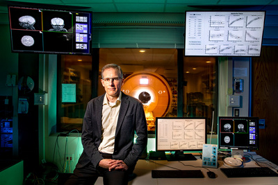 Researcher sits on a desk with readouts on computer monitors surrounding him and a magnetic resonance imaging device in the background.