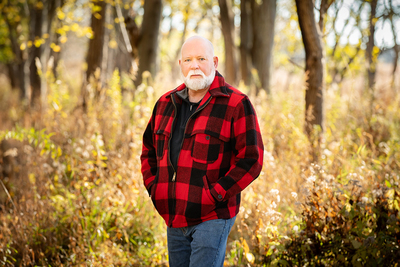 Craig Miller stands in the woods facing the camera. He is wearing a red and black plaid shirt and he has his hands in his pockets.