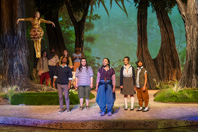 Photo of actors on stage with a woodland set and one character suspended above the others.