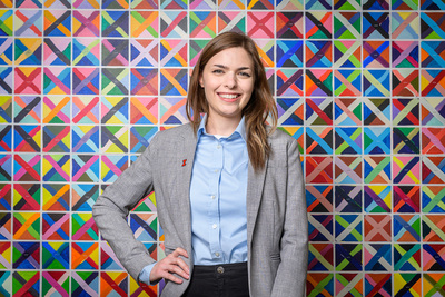 Educational psychology professor Kaylin Ratner wearing a blazer and button-down shirt, standing in front of a colorful mural with one hand on her hip