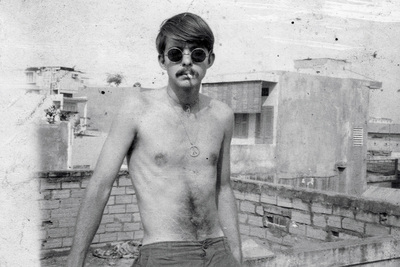 Black-and-white photo of a shirtless man wearing sunglasses and smoking a cigarette, leaning against a low wall on a rooftop with concrete buildings in the background.