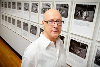 Photo of Tim Dean in a Krannert Art Museum gallery with Hal Fischer's photographic series "18th near Castro St. x 24" on the wall behind Dean.