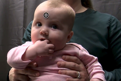 Photo of an infant in the IKIDS program seated on her mother’s lap. The infant has a sticker on her forehead that allows an eye-tracking instrument to orient to her eyes.