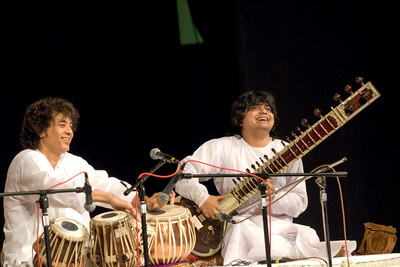 Photo of Zakir Hussain and Niladri Kumar, both dressed in white robes, sitting onstage. Hussain is playing the tabla and Kumar is holding a sitar.