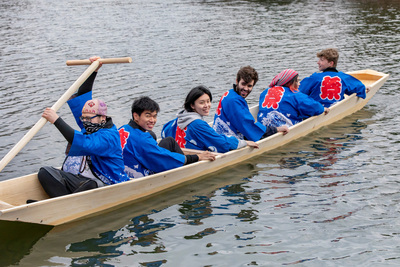 Photo of seven people in bright blue happi coats in a long, low Japanese riverboat on a pond.