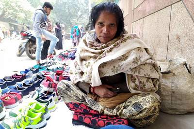 A female vendor in a mahila bazaar in New Delhi stares solemnly at the camera. A young man in the background is looking at the wares of a nearby vendor.