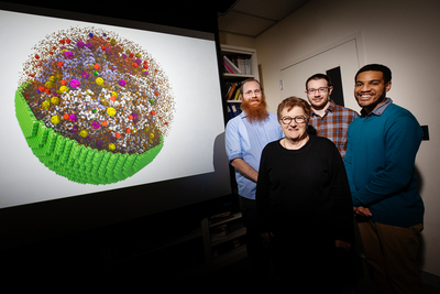 Research team stands in front of a screen with a 3D image of the minimal cell and its contents.