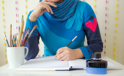 Person in blue sweater sits at desk with pens, paint brushes, and notebook ready to write something.