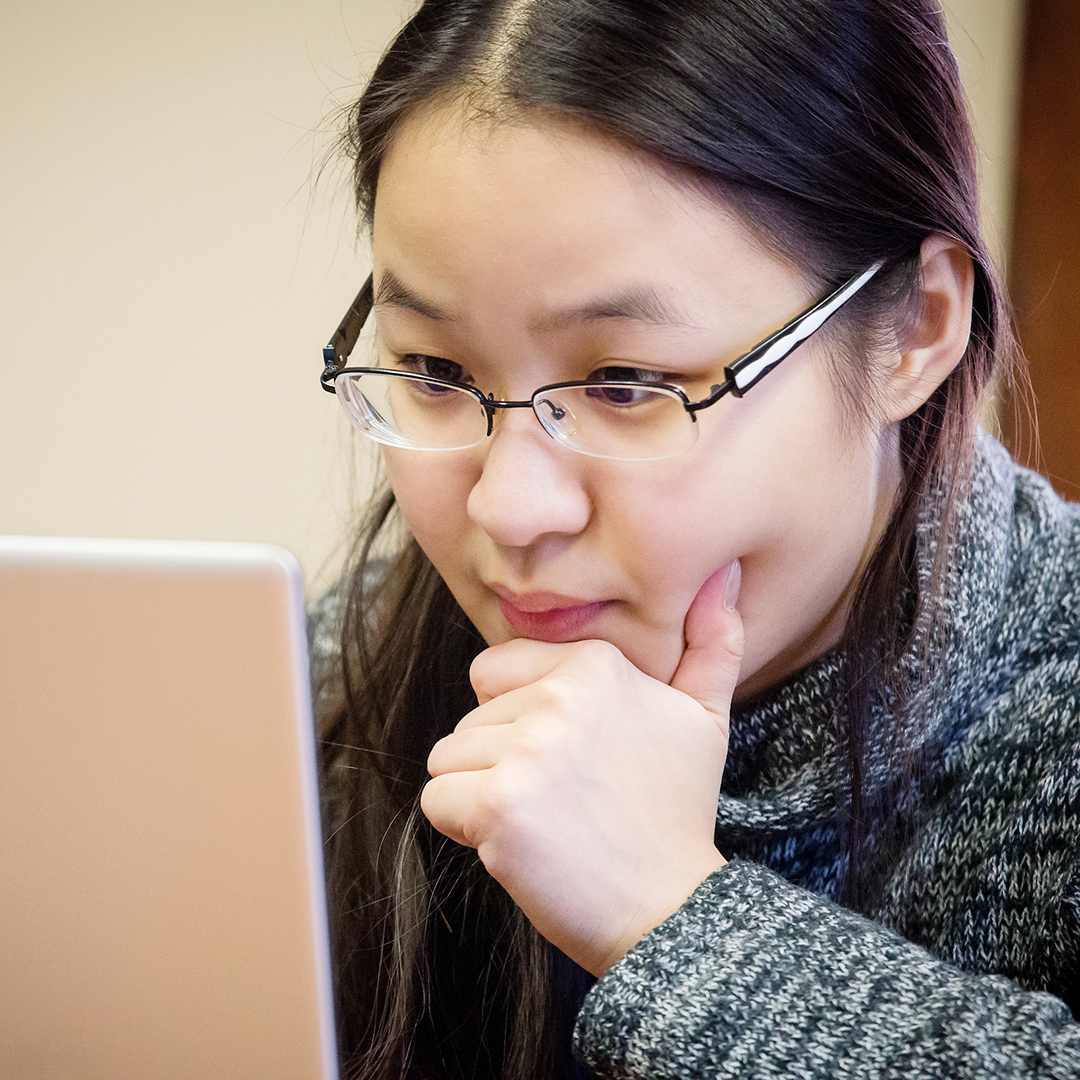 Asian woman wearing glasses, with her hand on her chin, looks quizzically at a laptop screen.
