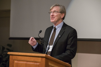 man at lectern smiling, gestures with one hand