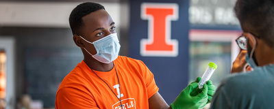 Masked Black male with orange SHIELD t-shirt h gloves handing test tube to man