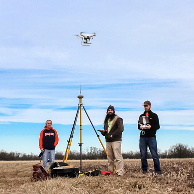 white drone hovering above 3 men in corn stubble field