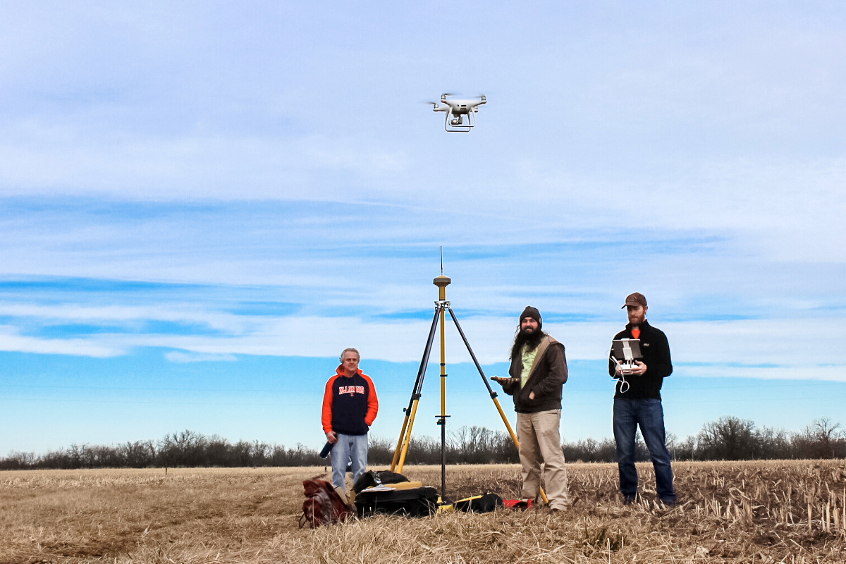 white drone hovering above 3 men in corn stubble field