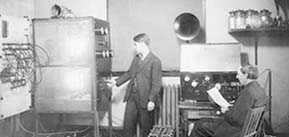 Two men in a room of early broadcasting equipment. One sitting holding a script near a microphone and the other standing near a machine adjusting dials.