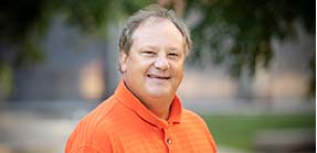 Headshot of Professor Stephen Moose wearing an orange U of I shirt standing in front of a blurred outdoor background