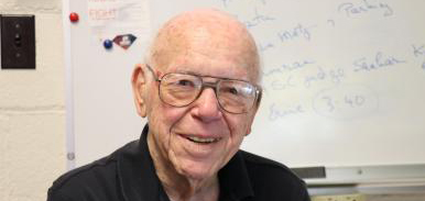 Harry H. Hilton, an elderly white man with a bald head and glasses, smiles at the camera. He is in front of a whiteboard with writing on it.