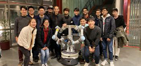 [Image ID: a group of 15 people clusters and poses around a silver and white robot, that has a cyllindrical body and two arms. End ID]