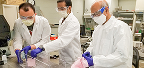 Three people in lab coats, goggles, gloves, and face masks hold squares of fabric in a clear plastic trough and spray them.