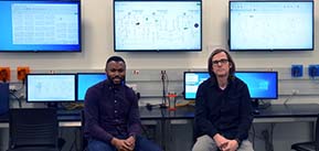 Research Engineer Olaolu Ajala (left) and Professor Alejandro Dominguez-Garcia (right) sitting in front lab equipement with their research displayed faintly on computer screens above and behind them.