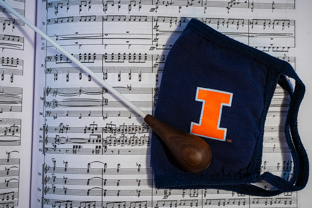 A baton and dark blue facemask with an orange I laid on top of a musical score.