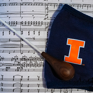 A baton and dark blue facemask with an orange I laid on top of a musical score.